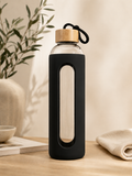 Black 1L glass water bottle with protective sleeve, bamboo lid and carry loop, styled in a warm minimalist wellness setting.