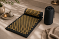 Black acupressure mat with gold pressure points, matching pillow and carry bag in a calming neutral home wellness setting.